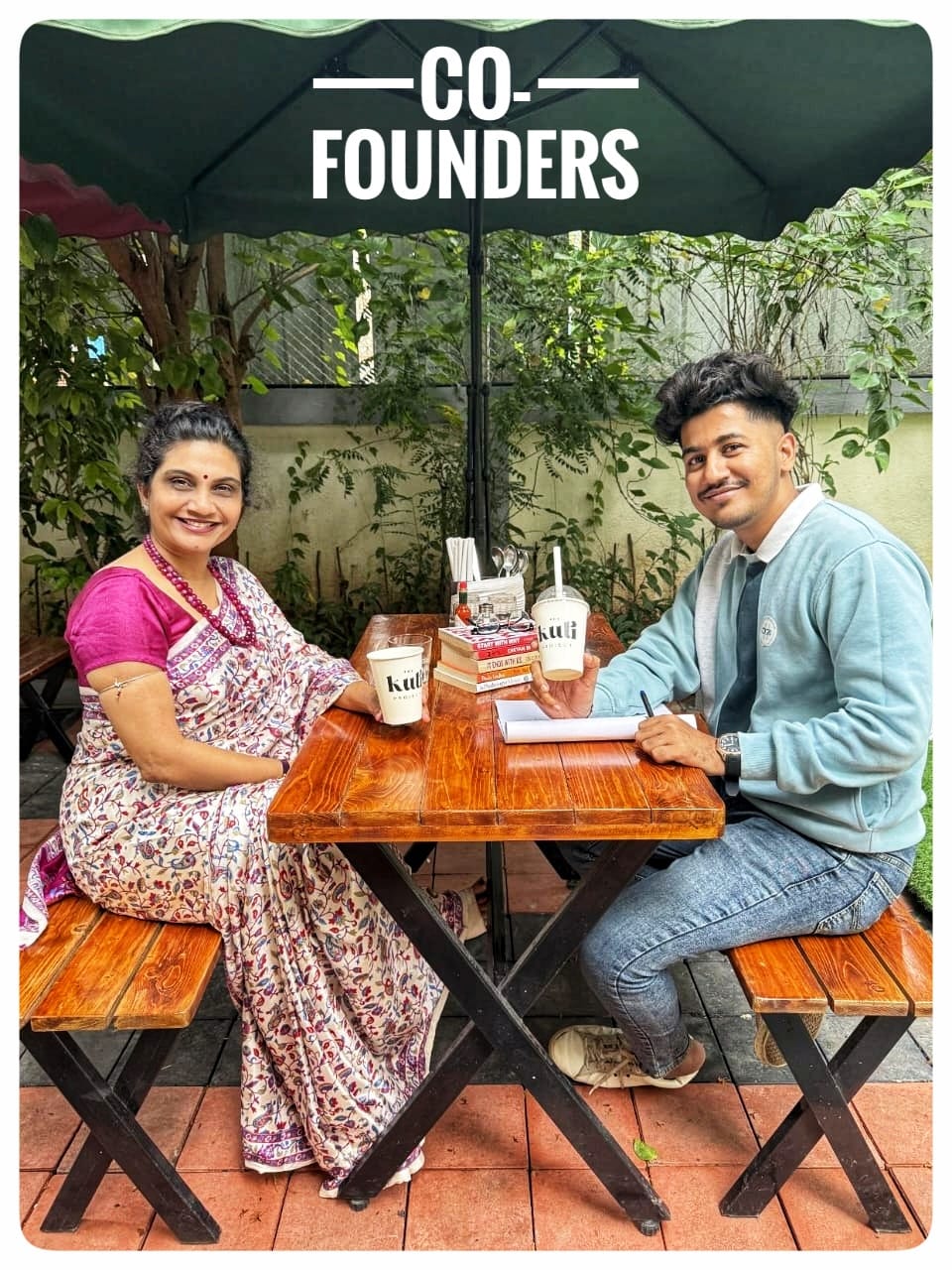 Two people that is Dr.Dipti Tulpule and Abhishek Prithviraj are sitting at a table under a 'Co-Founders' umbrella in an outdoor setting.